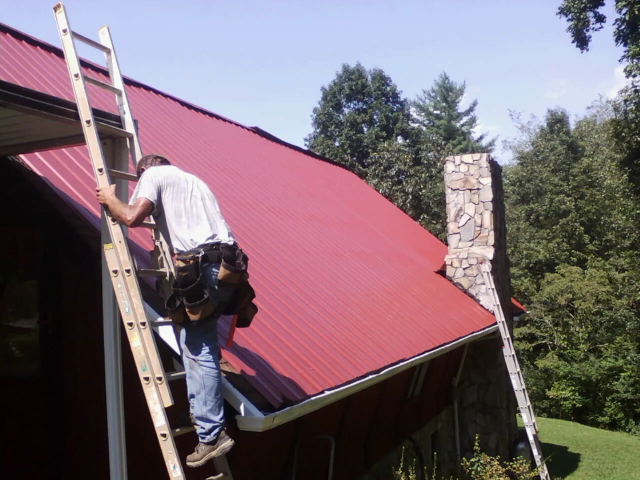 Roofer on a ladder beside a red metal roof and stone chimney — Soffit & Fascia Repair by Roofing Brothers in Creekside